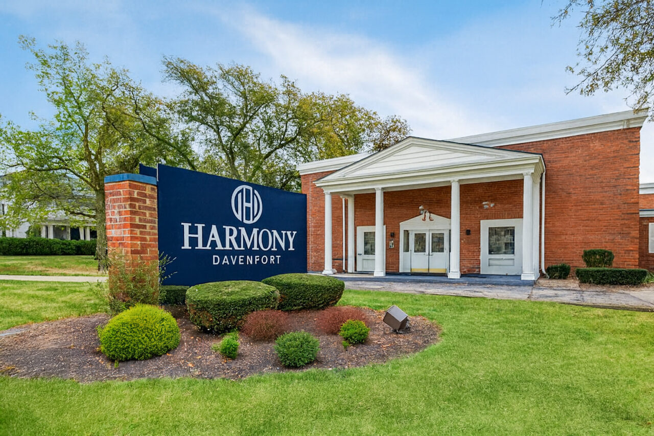 A brick building with a white portico entrance supported by columns, surrounded by a well-maintained lawn and small bushes. In front, there is a large dark blue sign with white text that reads 