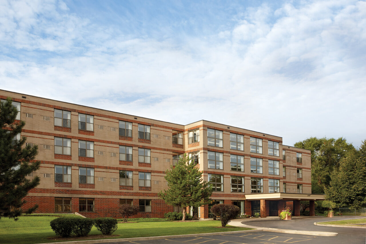 A multi-story beige brick apartment building with large windows, surrounded by green trees and bushes, under a partly cloudy blue sky.