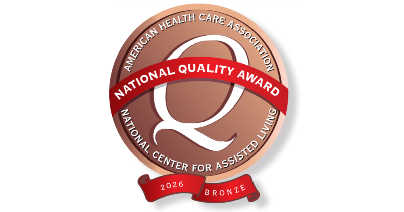 A circular badge with a bronze background, surrounded by a dark border. At the top, it reads "AMERICAN HEALTH CARE ASSOCIATION" and at the bottom, "NATIONAL CENTER FOR ASSISTED LIVING." A red ribbon across the center states "NATIONAL QUALITY AWARD." Below the ribbon, a smaller red banner displays "2026 BRONZE." The badge features a large, stylized "Q" in the center.