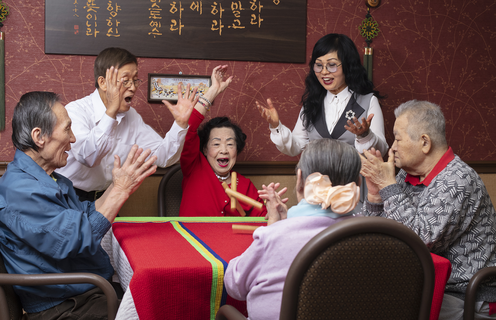 A group of elderly people and a woman are celebrating and playing a game at a table with a red, green, and yellow tablecloth. The woman in the center, wearing a red outfit, is smiling and raising her hand. The woman standing nearby, wearing glasses and a gray vest, is clapping and smiling. The other people are clapping, smiling, and engaging happily in the activity. The background features Korean calligraphy on the wall and traditional decorations.