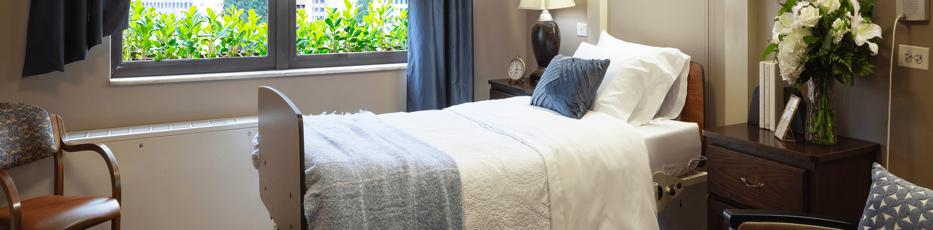 A hospital room with a bed covered in white linens, a gray blanket, and pillows, next to a nightstand with a lamp, clock, and flowers. A window with greenery outside is behind the bed, and there are chairs nearby.
