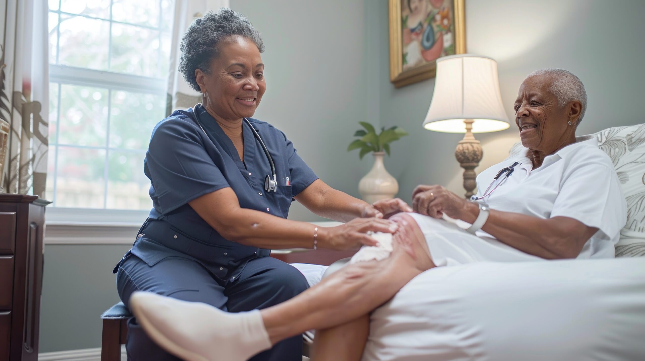 A healthcare worker in navy scrubs is assisting an elderly patient in bed, holding the patient's foot and smiling. The patient is lying on their back, smiling, with a stethoscope around their neck, in a well-lit room with a window, lamp, and artwork on the wall.