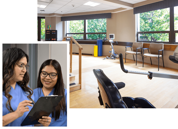 A therapy or rehabilitation room with large windows showing green trees outside, equipped with chairs, a stationary exercise bike, and padded foam rollers. In the foreground, two smiling female healthcare workers in blue scrubs are reviewing a clipboard together.