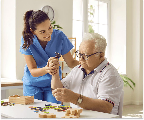 A young female caregiver in blue scrubs helps an elderly man with a wrist exercise, smiling and engaging with him at a table with game pieces and wooden blocks, in a bright room with large windows.