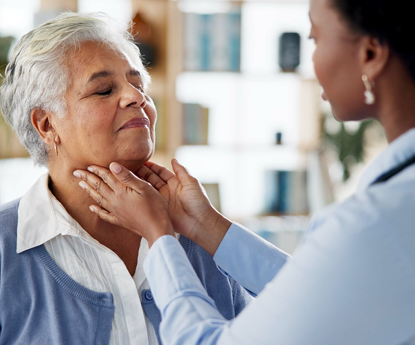 A healthcare worker gently checks an elderly woman's neck and throat area, who appears calm and content. The setting is indoors, possibly in a medical or home care environment.