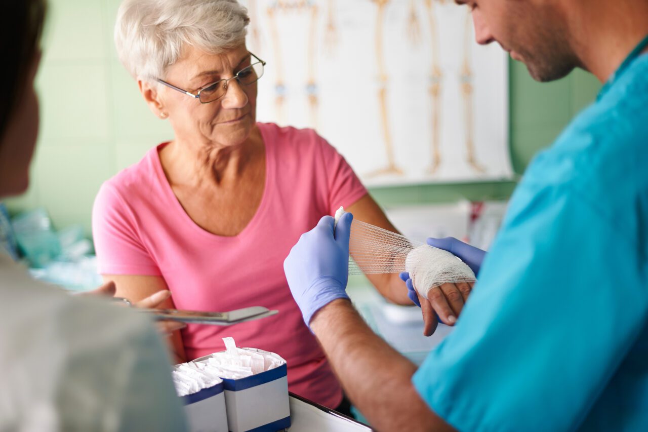 A healthcare professional applying a bandage to an elderly woman's hand, with a focus on wound care. The woman is wearing a pink shirt, and the professional is in blue scrubs and gloves. A diagram of human leg bones is visible in the background.