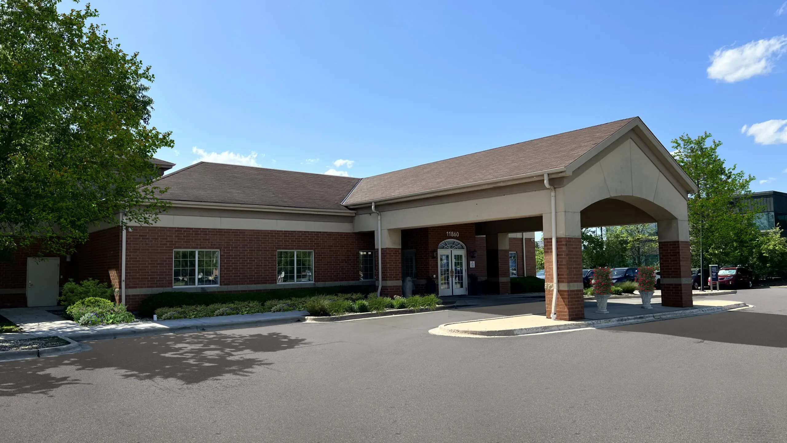 A brick building with a covered entrance supported by brick columns, surrounded by a parking lot with parked cars and landscaped plants, under a clear blue sky with a few clouds.